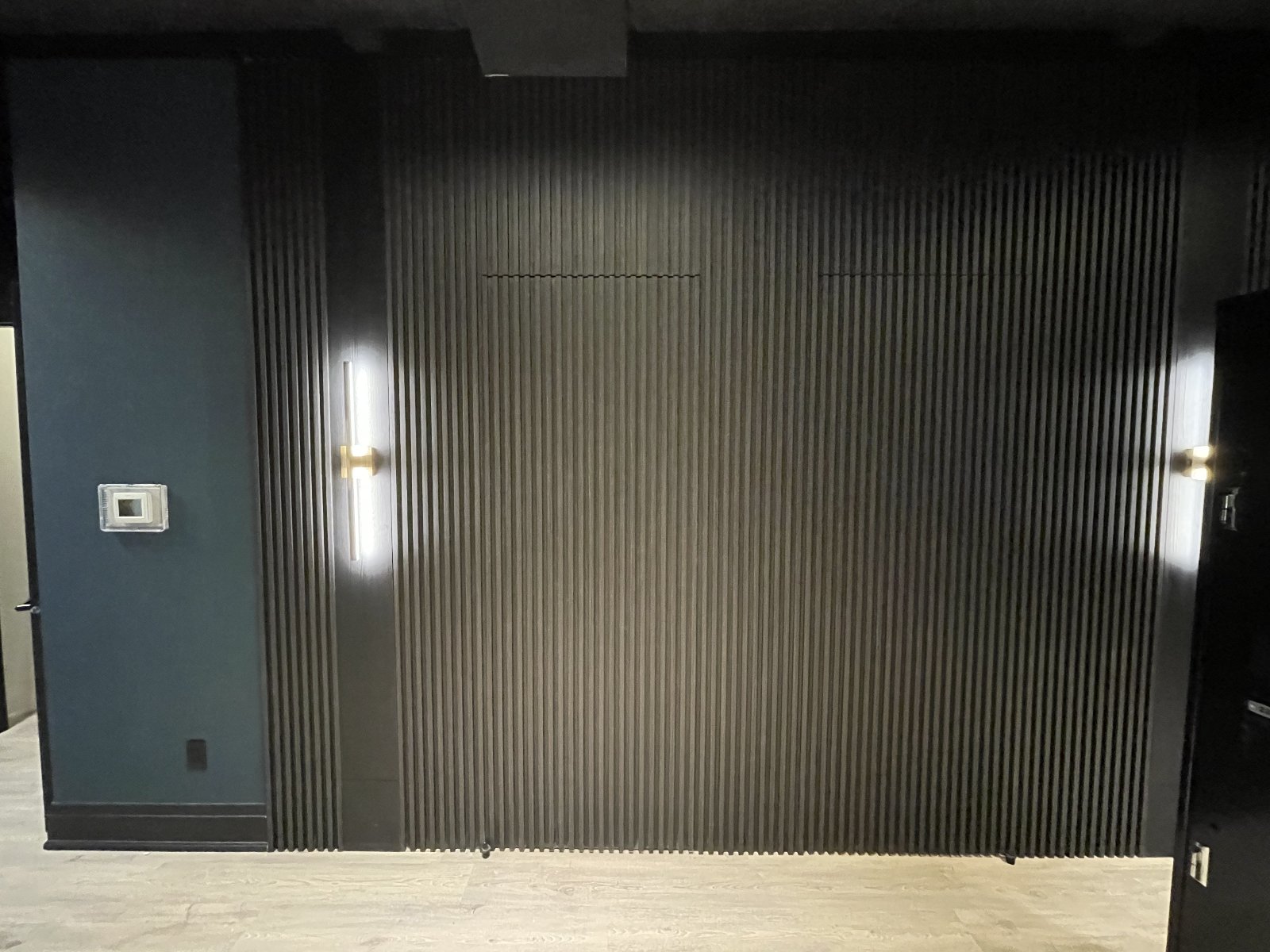 Feature wall detail — single brass linear sconce on the slatted backdrop, switched scene from the door.